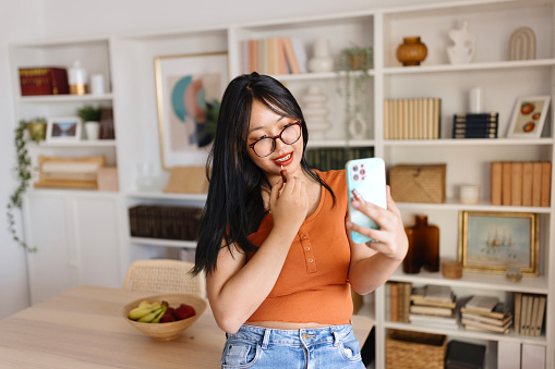 Young woman using her smartphone to put on her lipstick at home