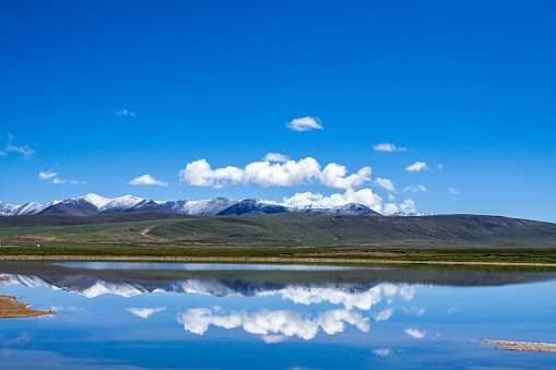 The water of the plateau lake reflects snowy mountains and grassland, blue sky and white clouds
