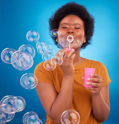 Fun, bubbles and black woman with joy, positive attitude and happiness against a blue studio background. African American female, lady and soap bubble for joy, playing and cheerful with excitement