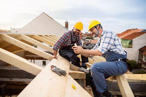 Two mid-adult male Caucasian roofers, taking an break from work while sitting on a roof beam, using mobile phone and drinking coffee or water