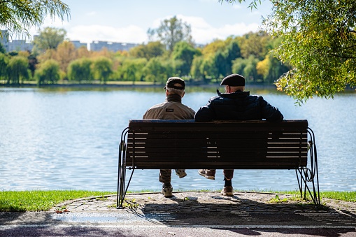 Bucharest, Romania – October 17, 2023: Two young Caucasian adult men sitting on a wooden bench outdoors enjoying the view of a tranquil lake
