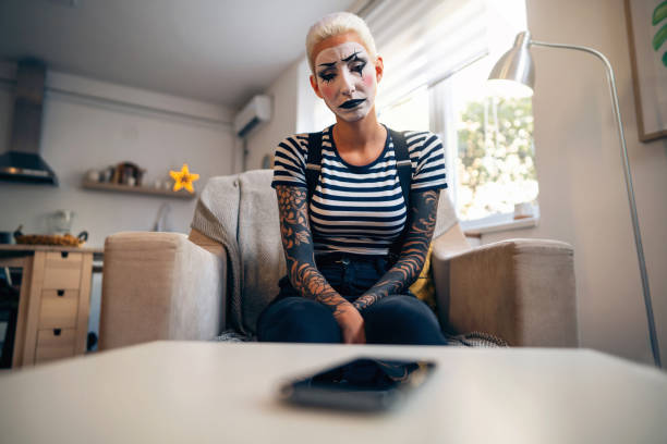 traurige junge frau, eine pantomime-künstlerin, die erwartungsvoll auf ihr handy schaut, während sie zu hause sitzt. - clown mime sadness depression stock-fotos und bilder