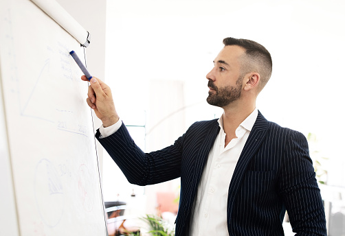 Serious confident caucasian mature man with beard in suit point at blackboard with graphs and charts in coworking office interior. Businessman, teacher, manager work, meeting and business planning