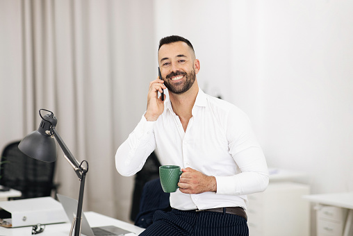 Happy confident european adult guy in white shirt with beard calls by phone, drinks cup of coffee in office coworking interior. Business, work and professional communication with client
