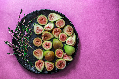 Still life with ripe sliced and whole fresh figs as healthy food for holidays. Black lava stone plate on purple concrete background and Heather as decoration. Rustic background. Copy space. Flat lay. Overhead view.