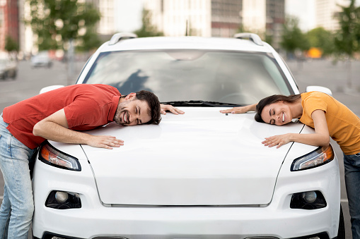 Happy young couple 30s handsome man and pretty woman hugging hood of their brand new white luxury car standing at parking spot outdoor. Transportation, auto leasing, buying, renting