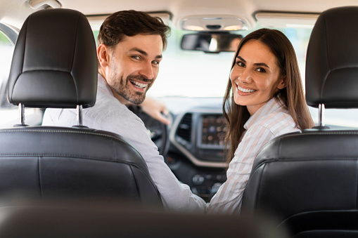 Joyful millennial spouses enjoying travel by car, sitting on front passengers seats in their new auto, looking at back seat and smiling. Caucasian man and woman driving and riding vehicle