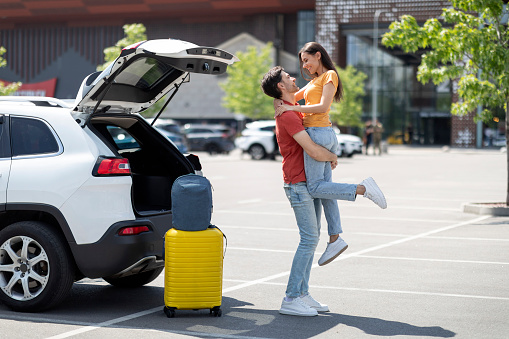 Happy loving millennial man lifting up his girlfriend, husband meet his beautiful wife at airport, back from journey. Emotional couple with luggage standing at parking outdoor by open car trunk
