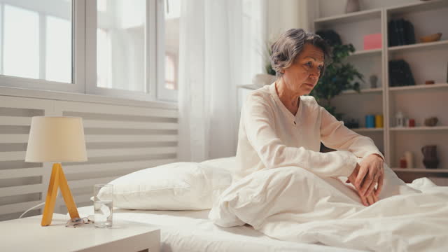Senior woman suffering from health disorder sitting on bed alone, feeling unwell