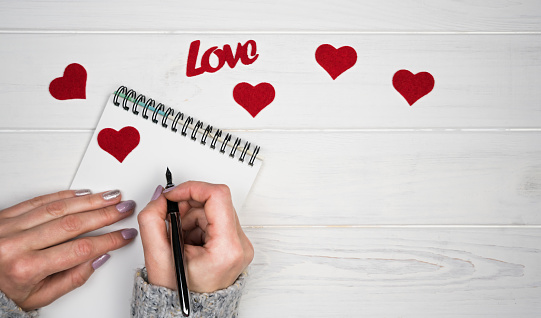 Woman's hands write a love letter on notepad. Romantic greeting for Valentine's Day. Valentine's Day holiday concept. Top view. Flatlay. Copy space.