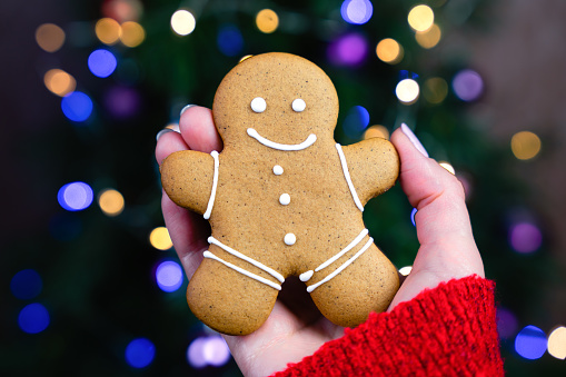 A woman's hand holds a gingerbread man on the background of the Christmas tree. DIY gifts. Festive mood. Close-up. Selective focus.