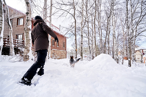 A man plays fetch with a dog in the snowy back yard of a house.