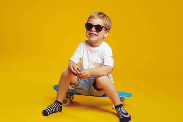 istock Cheerful kid in trendy sunglasses smiling and looking at camera while sitting on blue skateboard. 1782200959