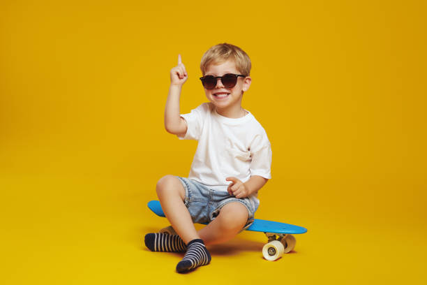 Excited kid wearing sunglasses and sitting on skateboard while looking at camera and pointing up.