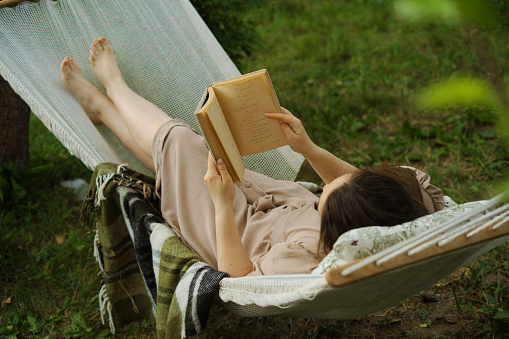 In a serene backyard, a woman finds escape in a book, swinging in a hammock. An embodiment of slow living and harmony, away from urban rush.