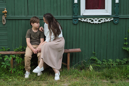moment of familial support captured, as a mother seeks to lift the spirits of her distressed elementary-aged son, both seated by their home, underlining the bonds of family.