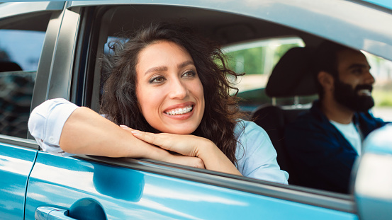 Portrait of smiling arab woman leaning out of car window and looking aside, happy lady sitting on passenger seat, enjoying automobile ride with her husband