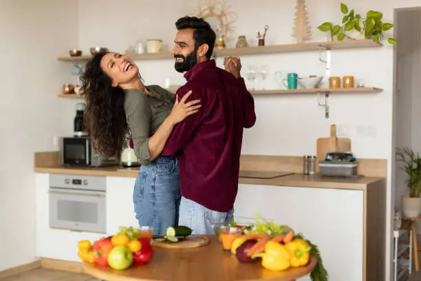 Excited arab spouses dancing together at kitchen and laughing, happy handsome man having romantic evening with his beautiful wife at home. Relationship, marriage, family concept