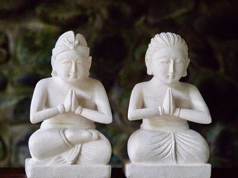 Horizontal closeup photo of two small stone sculptures sitting with legs folded in lotus position, hands together in prayer position, facing the front. Ubud, Bali.
