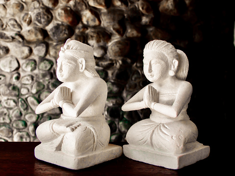 Horizontal closeup photo of two small stone sculptures sitting with legs folded in lotus position, hands together in prayer position on a wooden bench. Ubud, Bali.