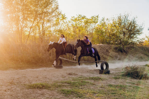 Two young women and their horses jump large jump while riding