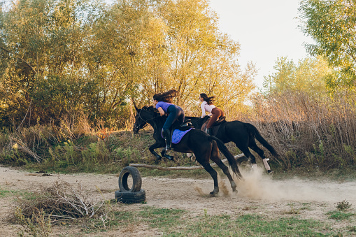 Two young women and their horses jump large jump while riding