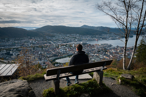 Traveler sitting on a bench, with his back turned, enjoying a panoramic view from a viewpoint of the city of Bergen, Norway.
