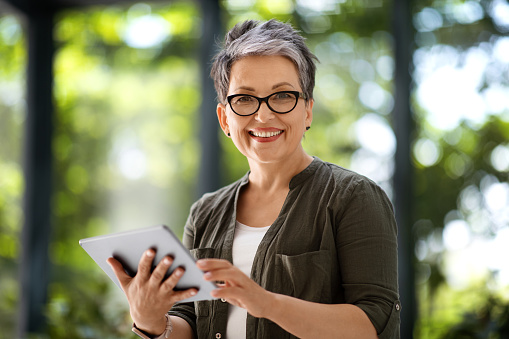 Happy attractive middle aged woman freelancer using digital tablet at home office, check job opportunities, blurred background. Corporate mature businesswoman websurfing, scrolling, check email on pad