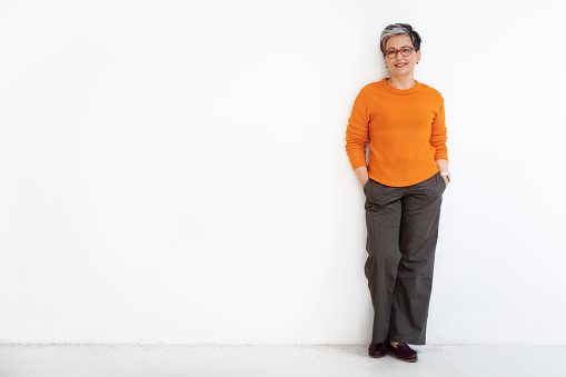 Happy beautiful stylish short-haired mature woman wearing casual outfit and eyeglasses posing with hands in pockets isolated on white background, copy space, full length. Boomers lifestyle