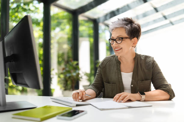 Attractive middle aged woman working on pc at green office Attractive cheerful short-haired middle aged woman wearing stylish casual outfit and eyeglasses IT project manager sitting at worplkace, working on pc computer at green office, working on report project manager remote stock pictures, royalty-free photos & images