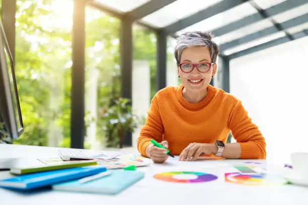 Attractive cheerful short-haired middle aged woman wearing stylish casual outfit, eyeglasses designer of interiors sitting at worplkace, working on pc computer at green office, using color paletter