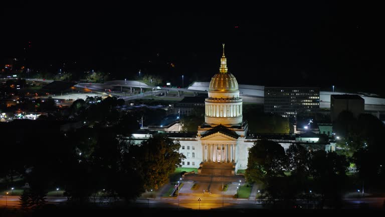 Backward Aerial Shot of West Virginia State Capitol Building with Charleston Behind at Night