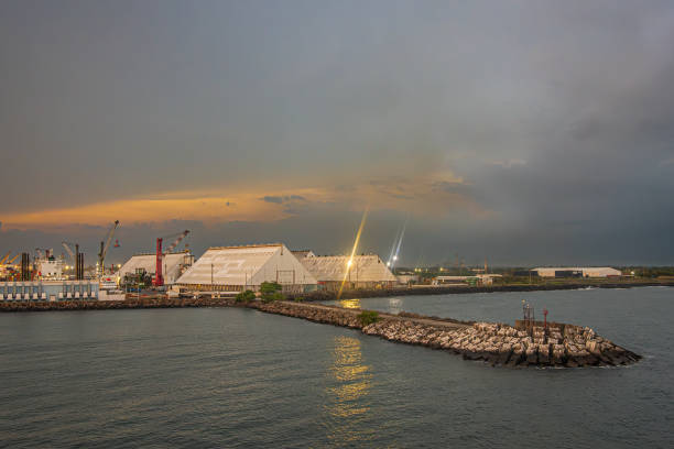 Guatemala, Puerto Quetzal - July 20, 2023: Evening cloudscape over tall white warehouses and cranes. Protective dam stretching onto Pacific ocean. Reflecting lights