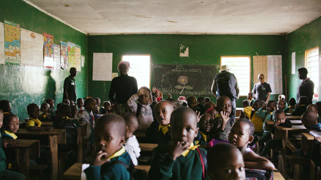 Slow motion shot of students waving to camera in classroom. Children with teacher and volunteers in school. Interior of educational building. Education is a basic human right that works to raise men and women out of poverty.
