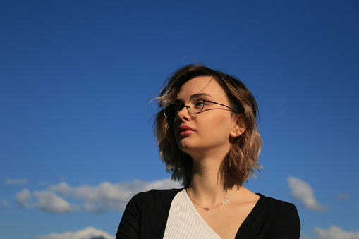 A headshot of a Russian female model on a sunny Autumn day. She is wearing short blond hair, glasses, makeup, jewelry and a black and white top.