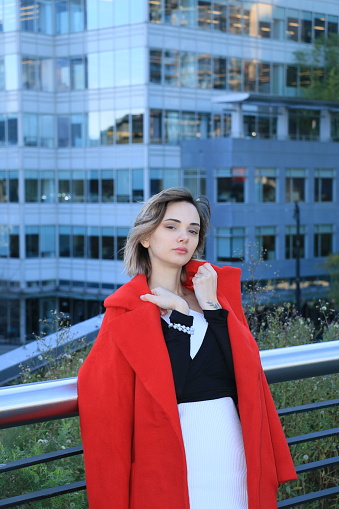A Russian woman leaning against a railing with a city building in the background. She is wearing short blond hair, makeup, jewelry, a red fully unbuttoned coat, black and white top, white skirt and tattoos.