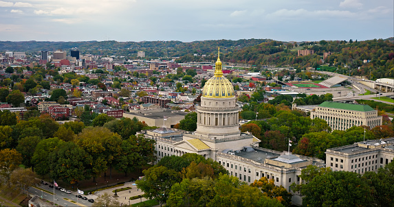 An aerial still image of the West Virginia State Capitol building with Charleston visible behind on a cloudy day.