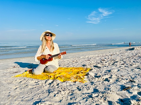 Active Senior  woman sits on yellow blanket playing ukulele on Indian Rocks Beach, Florida against water and blue sky