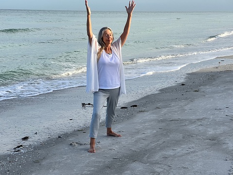Active senior woman reaches with arms above her head and she does Tai Chi on Florida Beavh against water and gentle waves