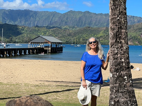 Senior woman stands on beach on the beautiful island of Kauai at Hanalei Bay near the pier against water and mountains.