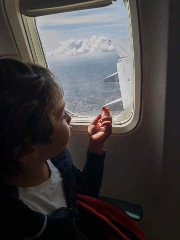 A little boy looking through the window of an airplane in flight. Bright sunny day with cloudy blue sky. He points his finger in the direction of the wing