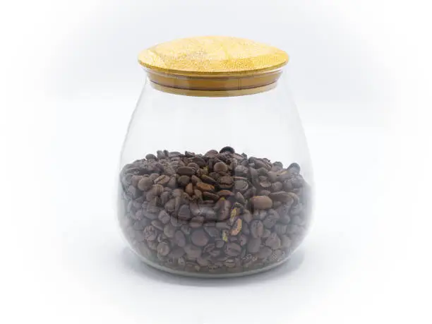 Photo of whole dark roast brown coffee beans in round glass jar or container with wooden top isolated on white background. front side view. number one most popular morning drink in the world to give energy