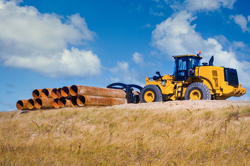 An yellow industrial tractor moves rusty pipes atop a grass covered sand dune on Cape Cod as part of a dune restoration project.