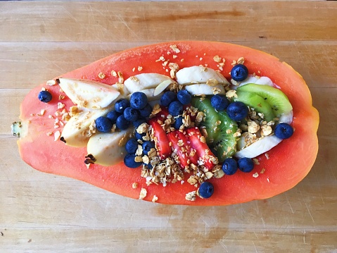 Top down close-up view of a papaya cut in half and filled with sliced fruit including kiwis, strawberries, guavas, blueberries and bananas and sprinkled with granola on a wooden cutting board