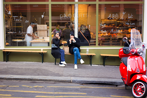 Paris, France: A young couple eating breakfast on a bench outside a bakery on Rue des Martyrs in the 9th arrondissement; a baker works inside.