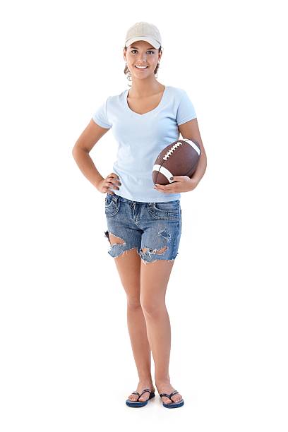 Sporty girl with American football smiling "Sporty girl holding American football, smiling, looking at camera..." woman wearing baseball cap stock pictures, royalty-free photos & images