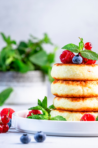 Thick pancakes with cottage cheese and blueberries, raspberries, red currants with fresh mint leaves on white plate. Gray table background