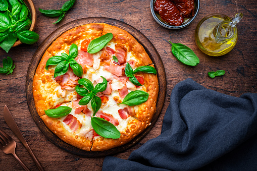 Homemade pizza with pancetta, mozzarella cheese, spicy sauce and green basil leaves on old wooden table background, top view