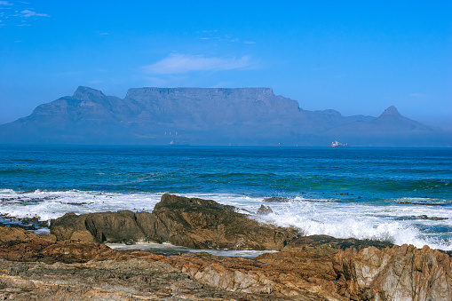 The rocky shoreline near Milnerton Beach in Cape Town, South Africa. In the far background is Table Mountain almost covered in fog. The image was shot in the afternoon sunlight. Horizontal format. No people.