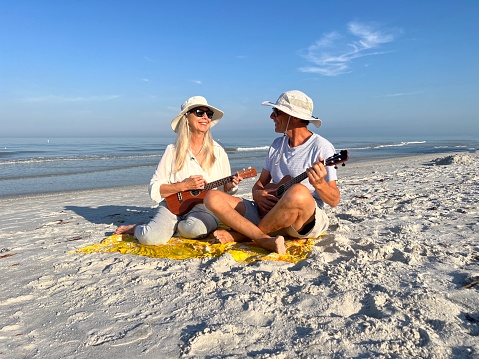 Active senior couple in sun hat and sunglasses  smiling at one another as they enjoy playing ukulele’s together as they sit on  Florida Beach against water and blue sky in the early morning